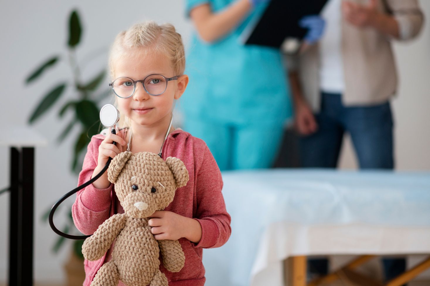 Ein Kind mit einem gehäkelten Teddy in der Hand trägt einen roten Pullover und hält ein Stethoskop. Im Hintergrund sind zwei Personen zu sehen.