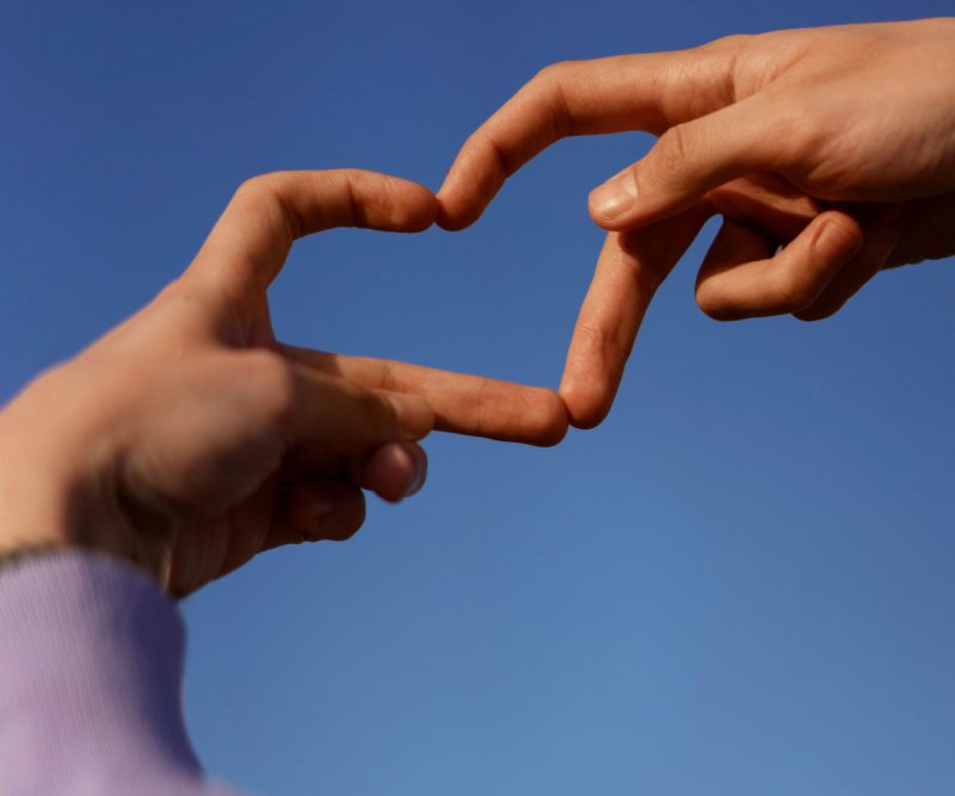 Zwei Hände formen ein Herzsymbol, die Finger sind leicht gekrümmt und berühren sich. Der Hintergrund ist blau.