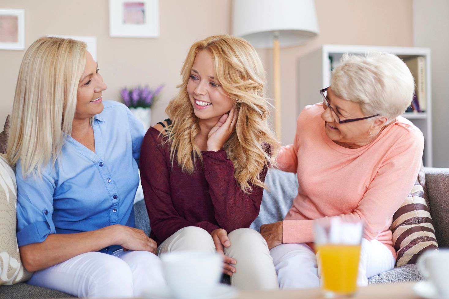 Drei Frauen sitzen auf einem Sofa. Die Frau links trägt ein blaues Hemd, die Frau in der Mitte hat lange blonde Haare und trägt einen roten Pullover, die Frau rechts trägt einen rosa Pullover. Auf dem Tisch steht ein Glas mit orangefarbener Flüssigkeit.